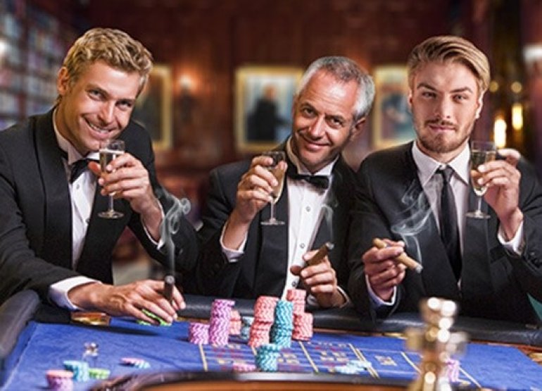 Трое мужчин в строгих костюмах пьют шампанское и курят сигары за игорным столом для рулетки
