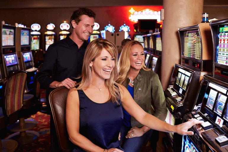 Две молоды женщины хорошо проводят время за игрой на автоматах в дорогой казино, а мужчина наблюдается за ставками, стоя на их спинами